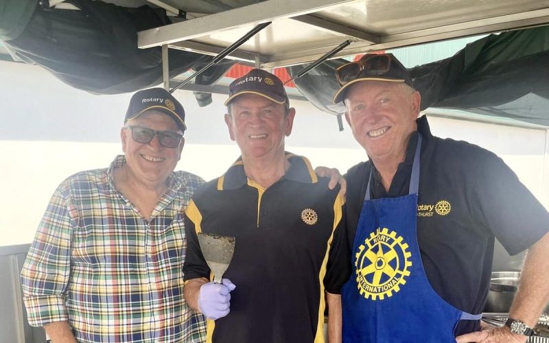Three club members in Rotary caps and apron smiling at the barbecue trailer.
