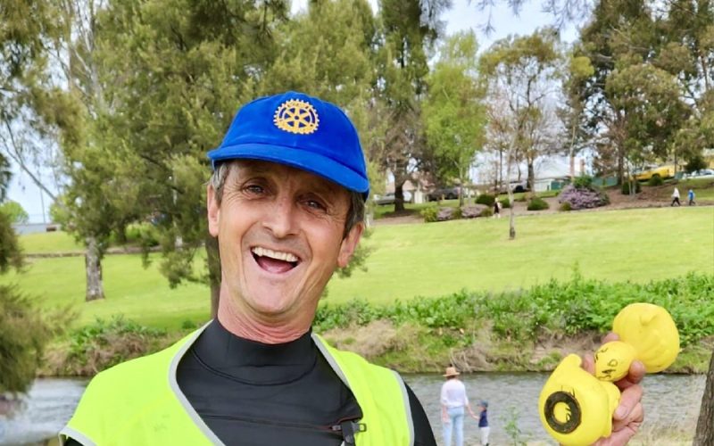 A club member in a hi-vis vest smiling, holding rubber ducks, at the riverside.
